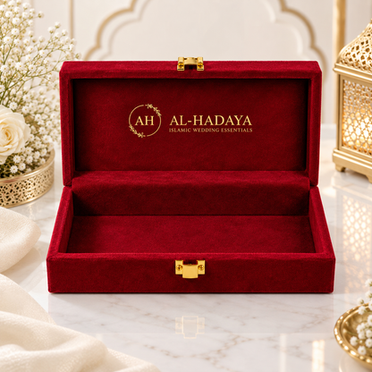 An open red velvet jewelry box with gold hardware, featuring the brand name 'AL-HADAYA' printed in gold on the inside lid, styled on a marble surface with decorative flowers and lanterns.