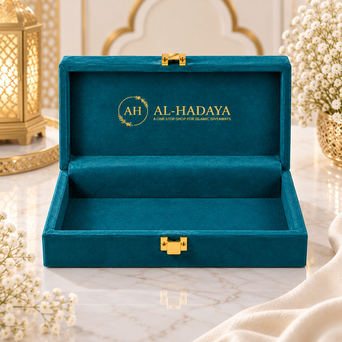 An open teal velvet jewelry box with gold-tone hardware, featuring the brand name 'AL-HADAYA' printed in gold on the inside lid, set on a marble surface with decorative flowers and a lantern in the background.