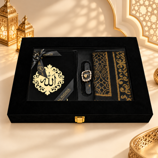 A luxury black gift set presented in a black velvet box with gold accents, featuring a decorative gold plaque with Arabic calligraphy, a black and gold patterned fabric item, and a black wristwatch with a crystal-embellished circular dial, styled on a warm beige surface with ornate gold lanterns in the background.