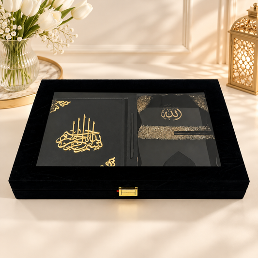 A black wooden keepsake box with a glass lid, shown open to reveal a black book and a black garment with gold Arabic calligraphy, set on a light cream surface with decorative flowers and a lantern in the background.