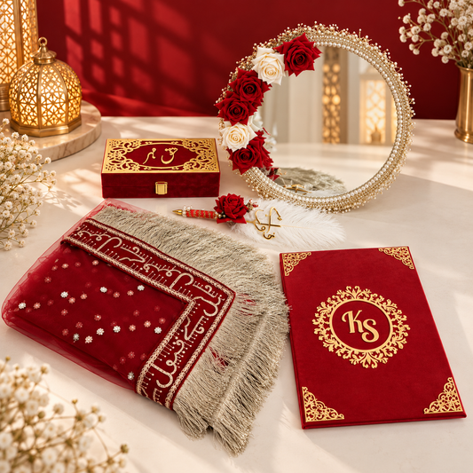 A styled flat-lay of a red and gold Islamic prayer rug alongside a matching red monogrammed prayer mat, a red velvet box with gold detailing, and a decorative mirror adorned with red and white roses on a white surface against a deep red background.