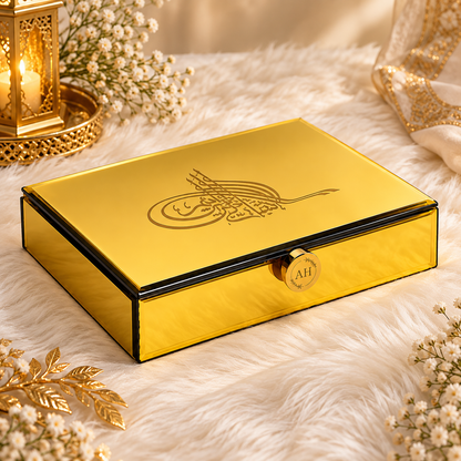 A shiny gold rectangular keepsake box with black trim and an engraved Arabic calligraphy design on the lid, set on a white faux fur surface with decorative gold lanterns and flowers.
