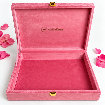 An open pink velvet jewelry box with gold-tone latches, shown on a white background with scattered pink flower petals. The inside lid is printed with the brand name 'AL-HADAYA'.