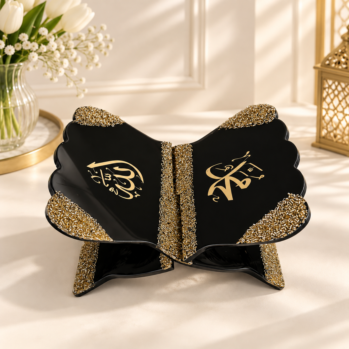 Black and gold decorative Islamic prayer mat with gold Arabic calligraphy on a cream-colored surface with soft shadows, styled with a vase of tulips and a lantern in the background.