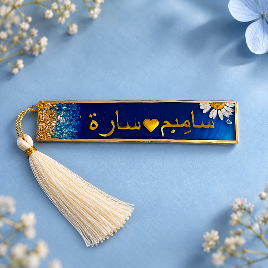 A rectangular enamel bookmark with a cream tassel, featuring gold Arabic calligraphy on a dark blue background with a glittery blue textured section on the left and a white daisy on the right. The bookmark is displayed on a light blue background with decorative baby's breath flowers.