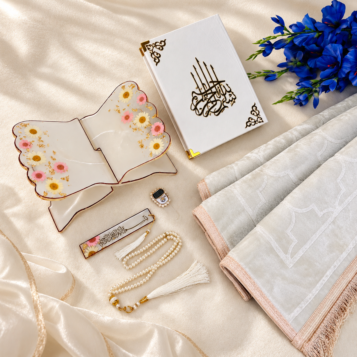 A styled flat-lay of a white Islamic prayer book with gold Arabic calligraphy, displayed with a white prayer mat, a decorative floral ceramic stand, a pearl tassel bookmark, a small prayer bead necklace, and blue flowers on a cream satin fabric background.