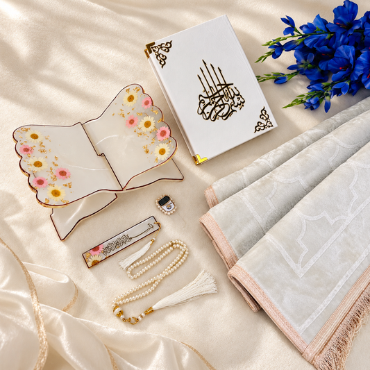A styled flat-lay of a white Islamic prayer book with gold Arabic calligraphy, displayed with a white prayer mat, a decorative floral ceramic stand, a pearl tassel bookmark, a small prayer bead necklace, and blue flowers on a cream satin fabric background.