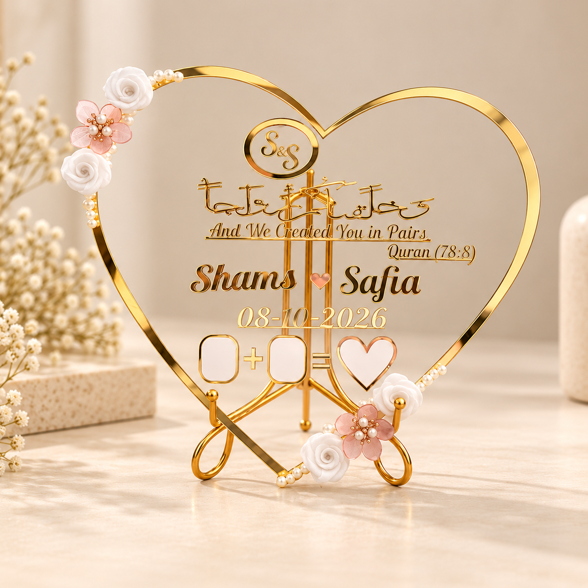 A gold heart-shaped acrylic and metal Islamic wedding plaque displayed on a light beige surface. The plaque features custom names 'Shams' and 'Safia', a date '08-10-2026', and placeholder boxes for dates and names, decorated with white and pink rose accents and pearl-like beads.