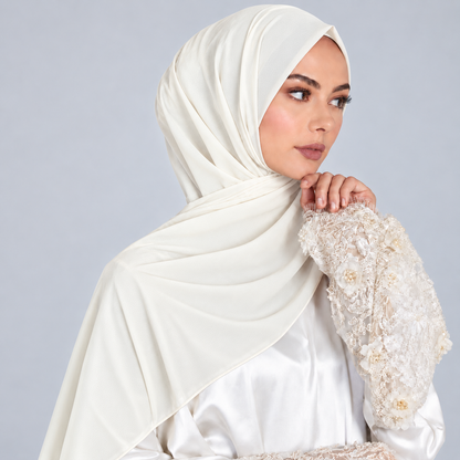 A woman wearing a draped off-white satin hijab and a matching embellished cream abaya, posed against a solid light gray studio background.