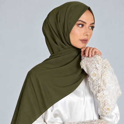 A woman wearing a draped olive green hijab and a white embroidered blouse, posed against a light gray studio background.