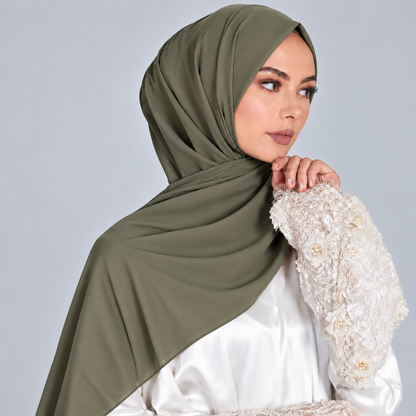 A woman wearing an olive green hijab draped elegantly over her shoulders and a white embellished blouse, posed against a solid light gray background.