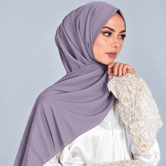 Elegant dusty lavender hijab worn in a graceful draped style with layered folds, paired with a white outfit featuring delicate floral lace detailing. Soft textured fabric, modest coverage, and refined feminine styling on a light blue-gray studio background.