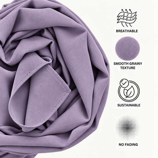 Elegant dusty lavender hijab worn in a graceful draped style with layered folds, paired with a white outfit featuring delicate floral lace detailing. Soft textured fabric, modest coverage, and refined feminine styling on a light blue-gray studio background.
