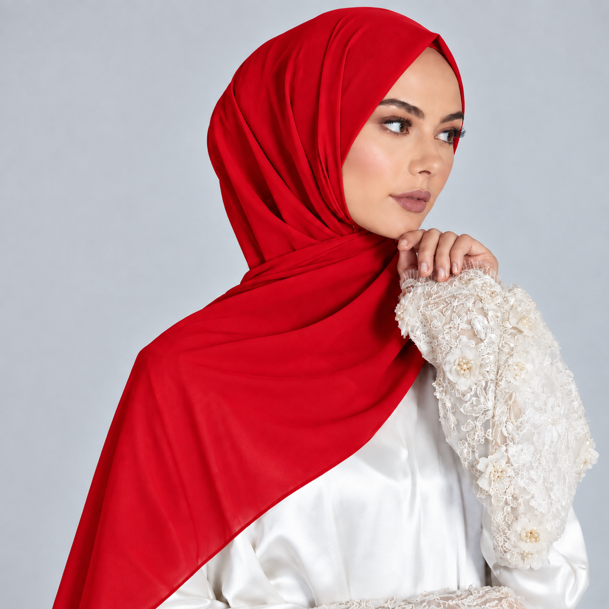 A woman wearing a bright red hijab and a white embroidered long-sleeve top, posed against a light gray studio background.