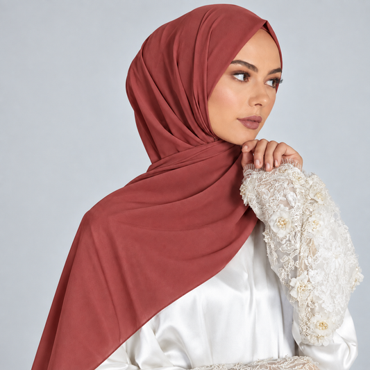 A woman wearing a draped dusty rose hijab and a white embroidered blouse, posed against a light gray studio background.
