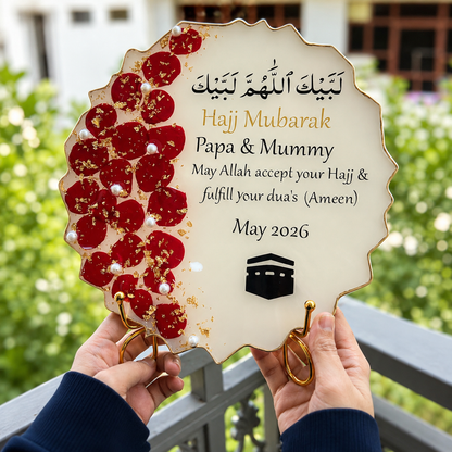 Hajj Mubarak stand with red floral design held by hands against a blurred green outdoor background