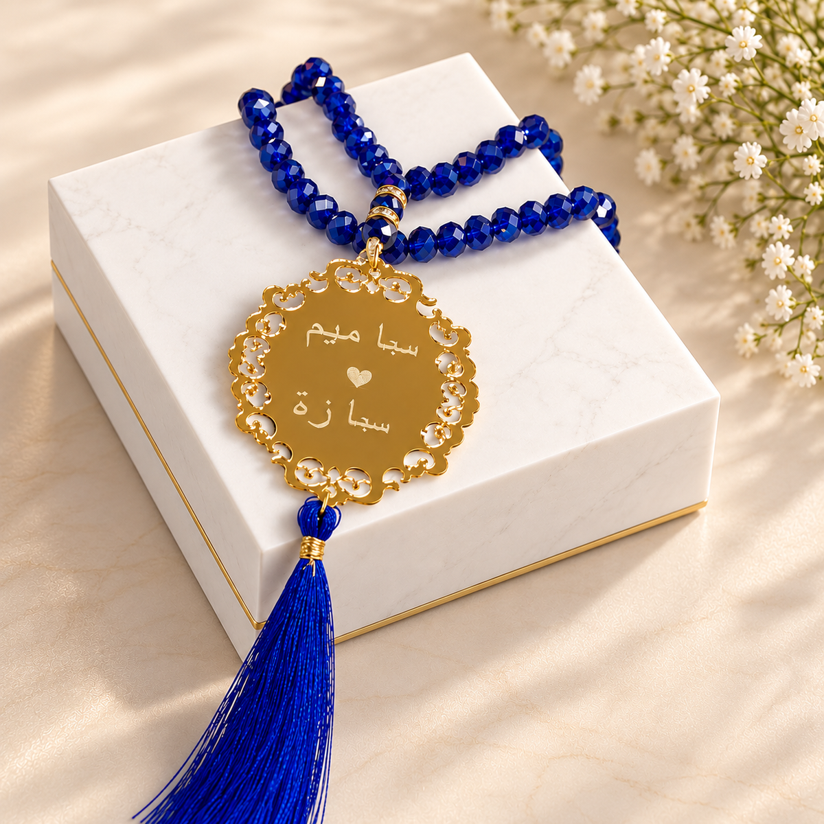 A blue beaded necklace with a gold-tone oval pendant and tassel, displayed on a white marble gift box with small white flowers in the background.