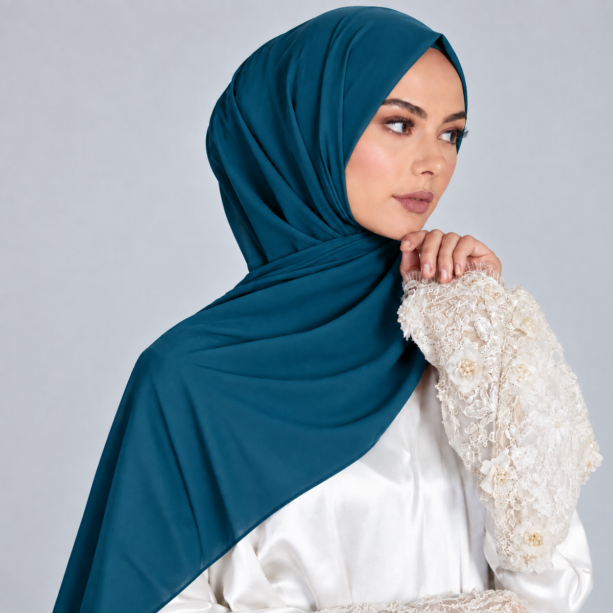 A woman wearing a teal hijab and a white embroidered blouse, posed against a light gray studio background.