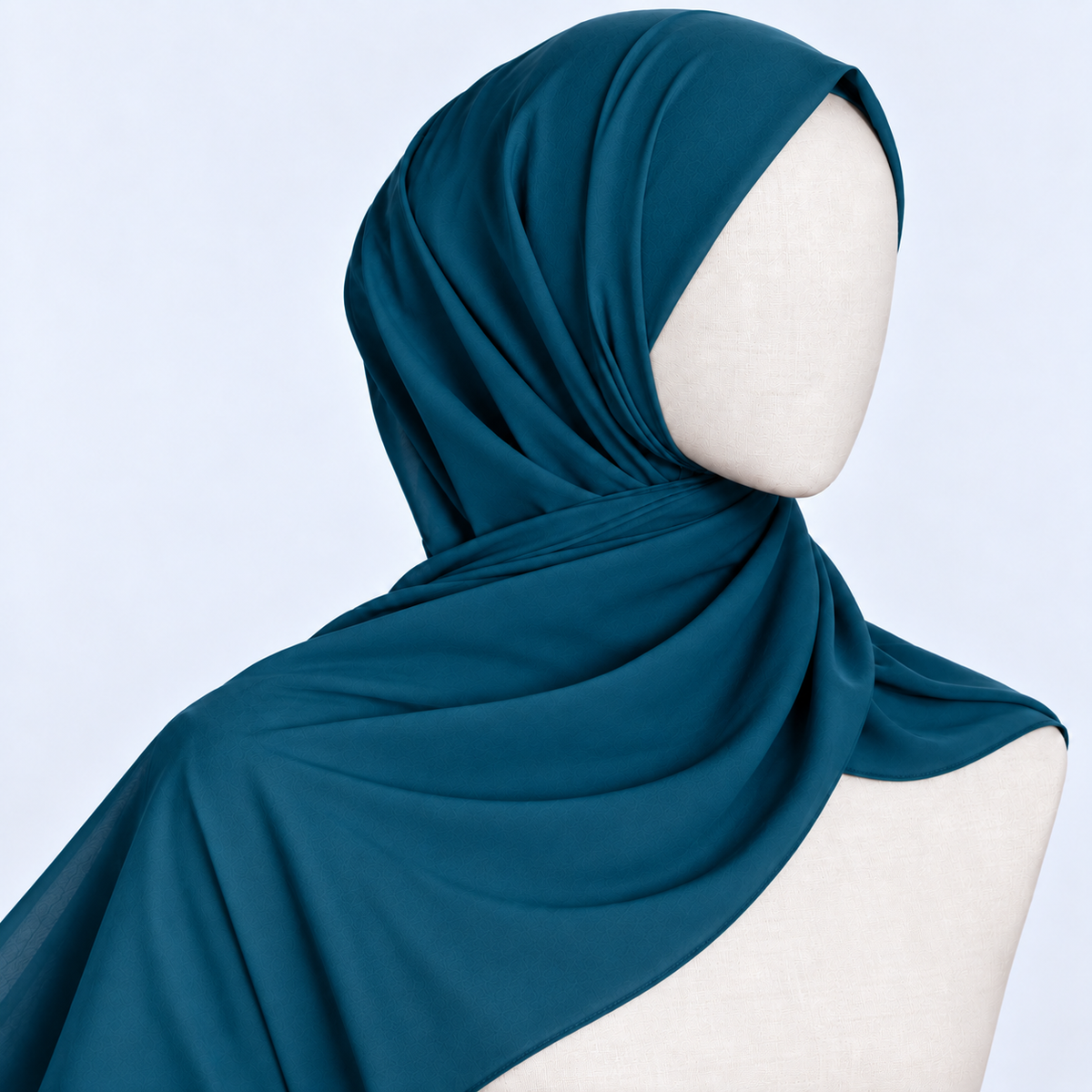 A dark teal blue hijab draped elegantly on a cream-colored mannequin head and shoulders against a light blue background.
