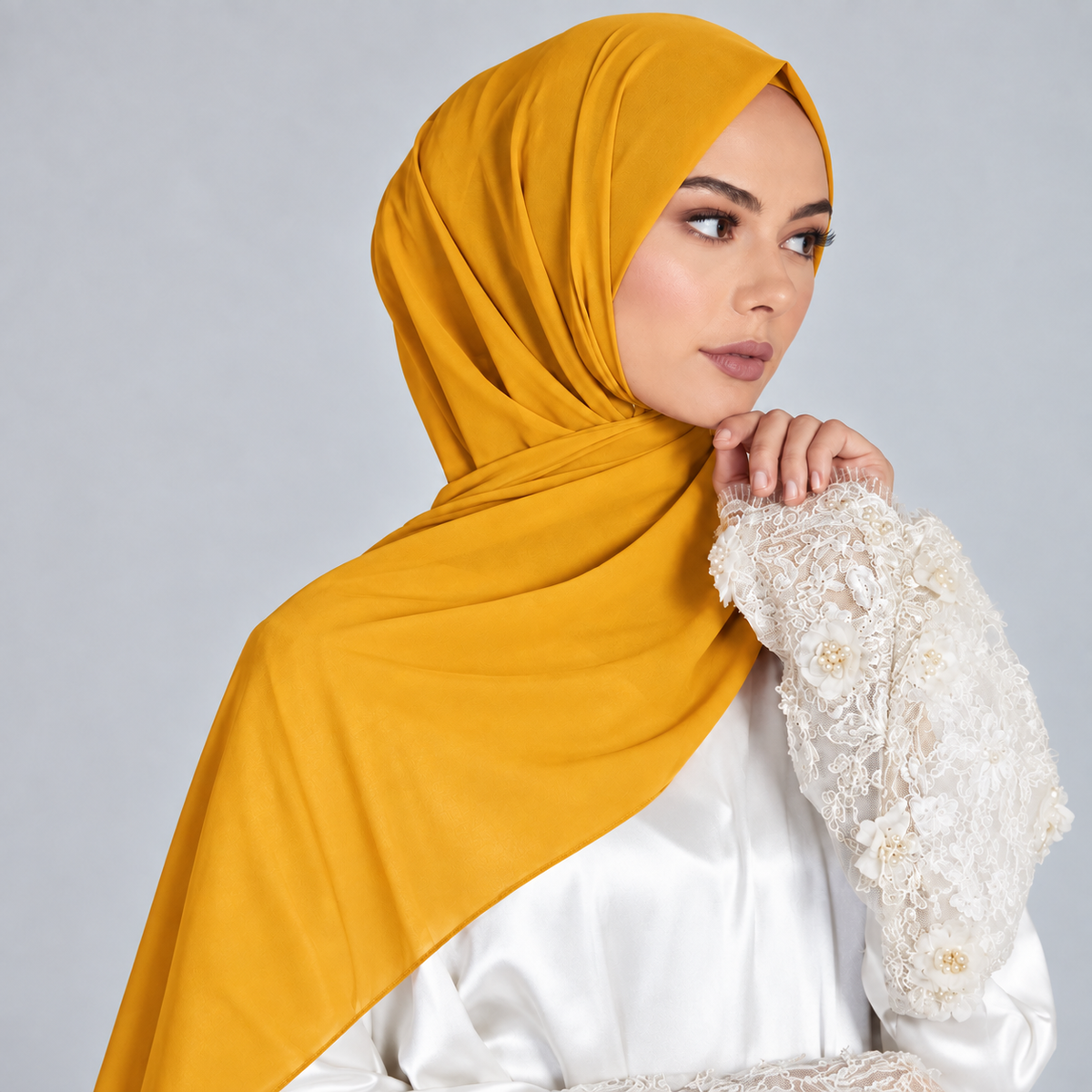 A woman wearing a mustard yellow hijab draped elegantly over her shoulders and a white long-sleeve blouse with intricate lace and gold floral embroidery on the sleeve, posed against a solid light gray studio background.